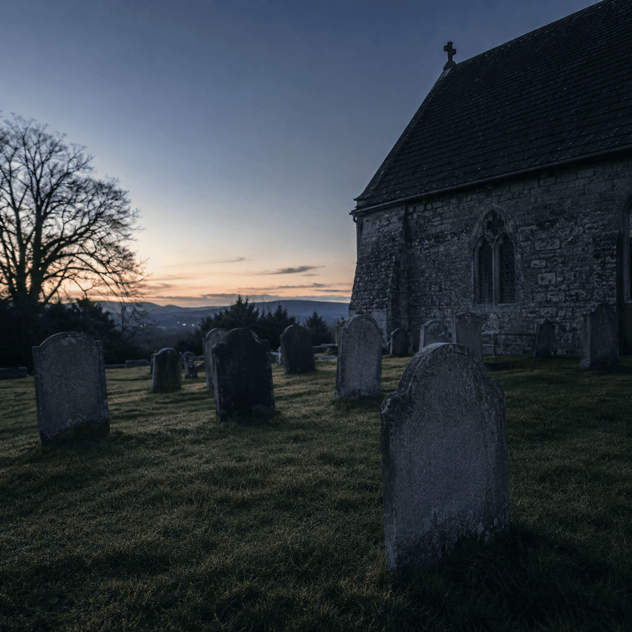 A quiet old country churchyard at dusk with weathered headstones beside a stone church