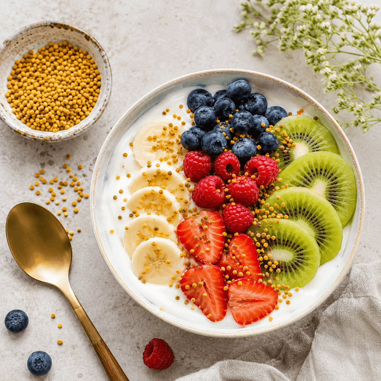 A breakfast yogurt bowl topped with bee pollen granules, sliced banana, blueberries, raspberries, strawberries, and kiwi
