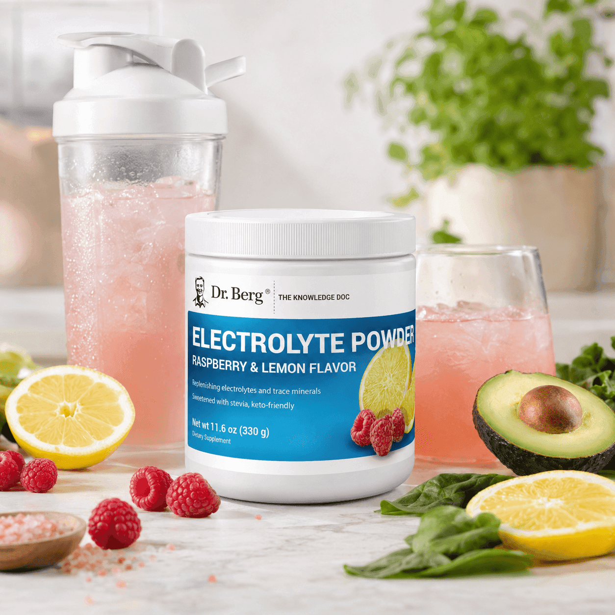 A tub of Dr. Berg Electrolyte Powder beside a filled shaker bottle and a glass of pink drink, surrounded by lemon, raspberries, avocado, and greens