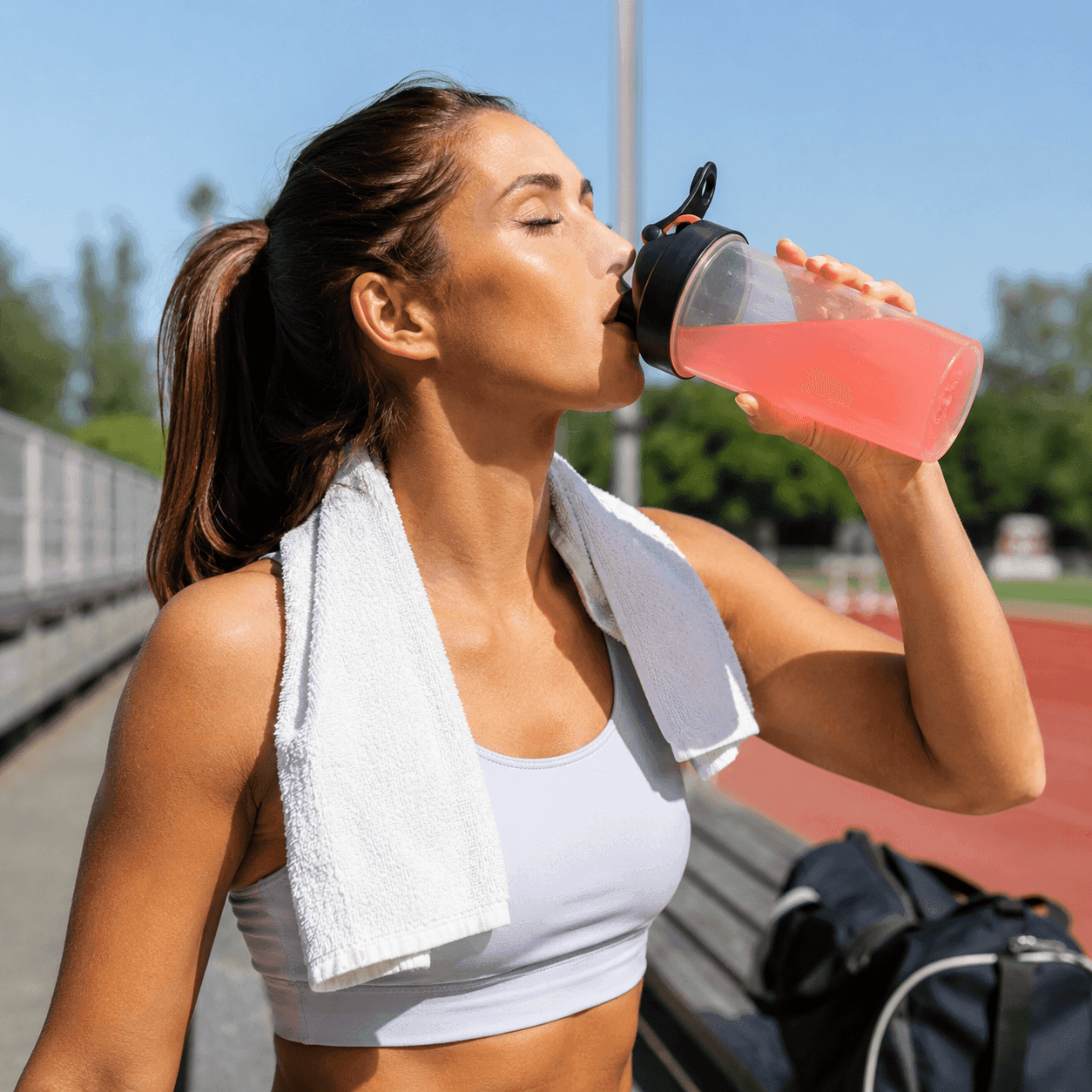 An athletic woman with a towel over her shoulder drinking a pink electrolyte drink from a shaker bottle at an outdoor running track